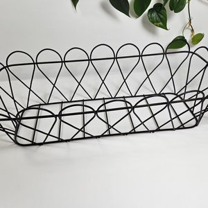 May include: Black metal wire basket with a scalloped edge design. The basket has a rectangular shape with a series of intersecting lines forming the base and sides. The basket is empty and sits on a white surface.