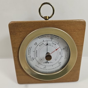 May include: A wooden barometer with a brass-colored bezel and a circular dial. The dial features a needle pointing to "Fair" and text indicating weather conditions like "Stormy" and "Dry". A brass ring is at the top for hanging.