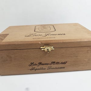 May include: A rectangular wooden box with a hinged lid and a gold-colored clasp. The box is light brown with the words "Leon Jimenez" and "Republica Dominicana" printed on it. The box is likely for cigars.