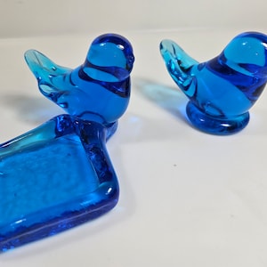 May include: Three pieces of blue glass bird figurines. Two birds are standing, and one is perched on a rectangular ashtray. The glass is a vibrant shade of blue, and the birds have a simple, elegant design. The ashtray is also blue.