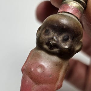 May include: A vintage glass Christmas ornament shaped like a person with a pink body and a brown head. The ornament has a metal screw top.
