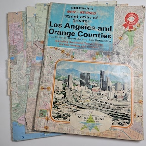 May include: A stack of vintage street atlases for Los Angeles and Orange Counties, featuring a cityscape illustration on the cover. The atlases include the text "GOUSHA'S NEW - REVISED street atlas of greater Los Angeles and Orange Counties".
