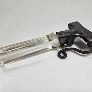 May include: A clear glass bottle opener with a metal handle and a hook. The handle is black and has a rusty appearance.
