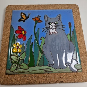 May include: A ceramic tile trivet with a cork backing features a gray cat with white markings sitting in a garden with green grass, red, yellow, and orange flowers, and a blue butterfly.