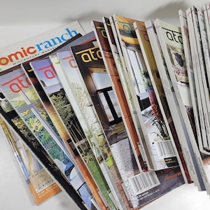 May include: A stack of "atomic ranch" magazines with colorful covers. The magazines feature images of mid-century modern homes and interiors. The magazine titles are in red and white. The magazines are stacked at an angle.