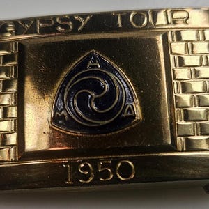 May include: A gold-colored belt buckle with the words "GYPSY TOUR" at the top and the year "1950" at the bottom. The center features a dark blue triangle with a swirling design and the letters "A" and "M". The buckle has a brick-like border.