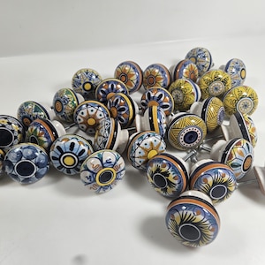 May include: A collection of colorful ceramic cabinet knobs with various floral and geometric patterns. The knobs feature a mix of blue, yellow, orange, and green designs, each with a unique pattern. The knobs are mounted on metal screws.