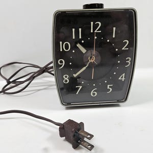 May include: A black vintage electric alarm clock with a silver trim. The clock face has white numbers and dots, with gold-colored hands. A black knob is on top, and a brown cord with a plug extends from the clock.