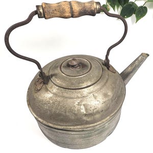 May include: A vintage metal tea kettle with a weathered silver-toned finish and a rounded body. It features a curved metal handle topped with a textured, light brown wooden grip and a matching circular lid with a small central knob.