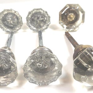 May include: A set of three clear glass door knobs with faceted, star-patterned centers. Two knobs feature long, slender silver-toned metal spindles, while the third knob has a shorter, darker, weathered metal spindle attached to its base.