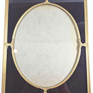 May include: A rectangular gold-toned metal picture frame featuring an oval inner border. The frame contains a dark black background matting and a central light-colored oval insert. Decorative metal accents are positioned at the top, bottom, and sides.