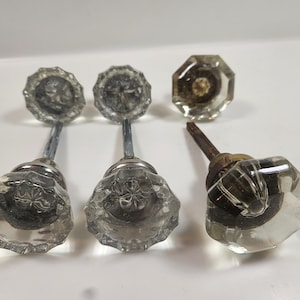May include: A collection of vintage glass door knobs. Three knobs have a faceted, flower-like design and long metal stems. Two additional knobs have a different shape and stem. The knobs are clear and reflect light.