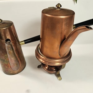 May include: Two copper coffee pots with black handles. One pot is taller and rests on a brass stand with a heating element. The other pot is shorter and has a tarnished appearance. Both have decorative finials on the lids.