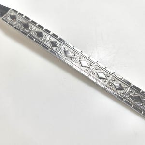 May include: A silver-toned metal fork handle featuring a repeating geometric pattern of raised diamonds set within square frames. The surface has a textured, stippled finish surrounding each diamond shape, creating a decorative border along the length.