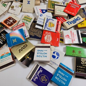 May include: A collection of vintage matchbooks with various designs and logos. The matchbooks feature a variety of colors, including red, blue, green, yellow, and white. Some of the matchbooks have text on them, such as "Holiday Inn", "Best Western", "Ramada Inns", and "Hilton Hotels".