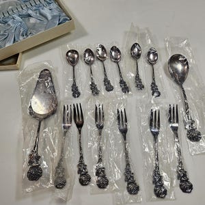 May include: A collection of vintage silverware, including spoons, forks, and serving utensils. The silver-toned pieces feature a floral rose design on the handles. Some pieces are individually wrapped in clear plastic, and a few are in a box with blue satin lining.