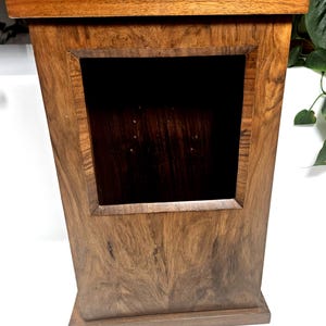 May include: A tall, rectangular wooden display case with a rich brown grain pattern. The structure features a wide top ledge, a solid base, and a large, square, open-front compartment in the center that reveals a dark, recessed interior space.