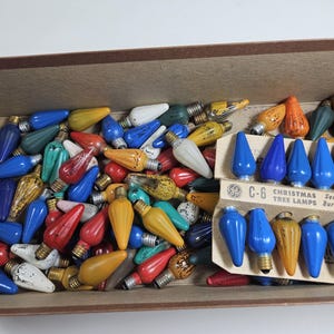 May include: A box filled with a variety of vintage C-6 series Christmas tree lights. The lights are in various colors, including red, blue, green, yellow, and white. The box also includes a card with the text "C-6 Christmas Tree Lamps Series Burning".
