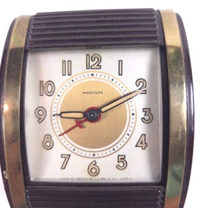 May include: A rectangular Westclox alarm clock featuring a dark brown ribbed casing with gold-tone metal side accents. The clock face has a white background, a central gold circle, black Arabic numerals, and black hour and minute hands with a red second hand.