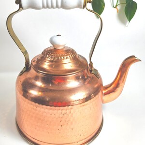 May include: A hammered copper tea kettle featuring a rounded body, a curved spout, and a brass handle topped with a white ceramic grip. The lid includes a matching white ceramic knob and decorative embossed detailing around the circular edge.