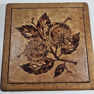 May include: A wooden framed tile with a brown leather surface. The tile features a woodburned design of two chestnut burrs with leaves.