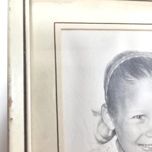 May include: A rectangular picture frame with a distressed off-white finish and a thin dark brown inner border. The frame contains a black and white portrait of a smiling child wearing a headband and a textured top, visible from the right side.