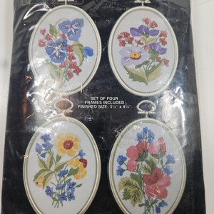 May include: A craft kit by Janlynn featuring four oval embroidery hoops with floral designs. Each frame contains a different arrangement of colorful flowers. The kit includes silk screened fabric, cotton floss, a needle, and instructions. The finished size is 3.5" x 4".