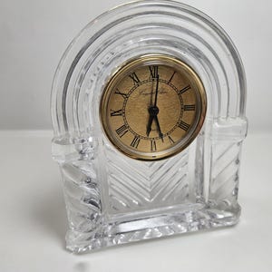 May include: A clear crystal clock with a gold-toned clock face and black hands. The clock has Roman numerals and the words "Crystal Clear" are printed on the face. The clock is in an arched shape.