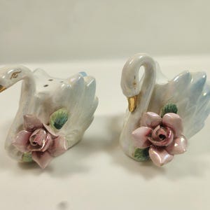 May include: A pair of ceramic swan salt and pepper shakers. The swans are white with light blue accents on their wings and gold beaks. Each swan has a pink rose at its base, with green leaves.