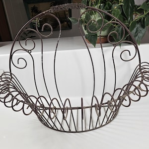 May include: A decorative, wrought iron basket with a handle. The basket has a curved design with scrollwork and vertical bars. The metal is a dark brown color, and the basket appears to be vintage or antique.
