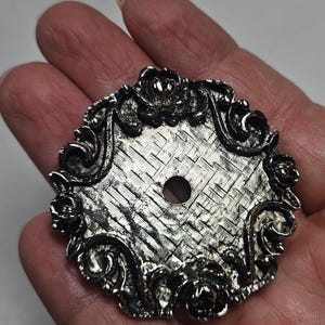 May include: Ornate, silver-toned metal decorative piece with a woven center and floral and scrollwork border. The circular design features a central hole and is held in a hand.