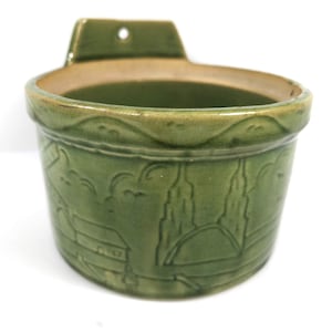 May include: A green ceramic wall pocket with a landscape design. The pocket has a rounded shape with a decorative rim and a small tab with a hole for hanging. The exterior features a relief pattern of buildings and trees.
