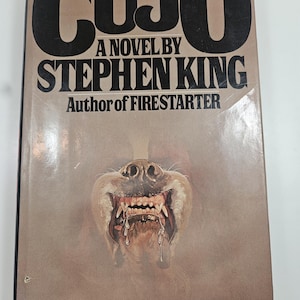 May include: Vintage paperback book cover for "Cujo" by Stephen King. The cover features the title in large, black, bold letters, with the author's name and a menacing illustration of a dog's snarling face with exposed teeth and saliva.