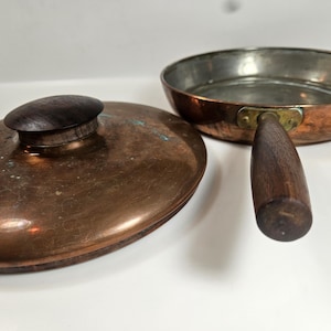 May include: A copper frying pan with a wooden handle and a copper lid with a wooden knob. The pan has a silver interior and a brass fitting where the handle is attached. The lid is oval-shaped.