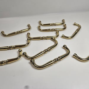 May include: A collection of nine brass-colored cabinet pulls. Each pull has a curved handle with decorative detailing and rounded ends. The pulls are arranged on a white surface, showcasing their design.