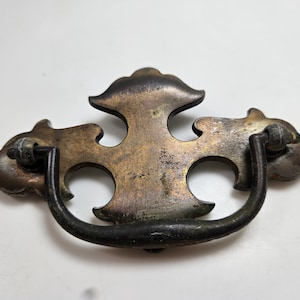May include: Antique brass drawer pull with a decorative design. The pull features a curved handle and an ornate backplate with a cross-like shape and two circular cutouts. The metal has a weathered, aged appearance.