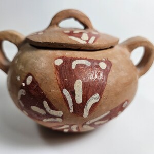May include: A small, brown ceramic pot with a lid. The pot has a white and red geometric design painted on it.