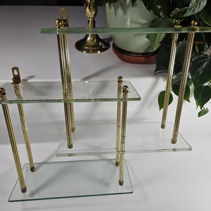 May include: A three-tiered glass shelf unit with gold metal supports. The shelves are clear glass and the supports are a shiny gold color.