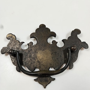 May include: Antique-style drawer pull with a dark bronze finish. The pull features an ornate, symmetrical design with a curved handle and decorative flourishes. The metal has a distressed appearance, adding to its vintage charm.