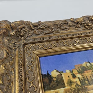 May include: A framed painting with a detailed, ornate gold-colored frame. The painting depicts a cityscape with buildings and a blue sky. The frame has intricate carvings and a distressed finish, adding to its vintage aesthetic.