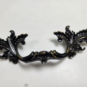 May include: Ornate, antique-style drawer pull with a dark bronze finish. The handle features intricate floral and leaf detailing, adding a touch of vintage elegance. Perfect for cabinets or furniture restoration projects.