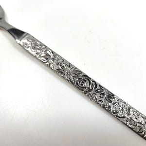 May include: A stainless steel fork featuring a flat, rectangular handle with an embossed floral pattern. The design includes detailed depictions of various flowers and leaves covering the entire length of the handle, ending in a straight edge.