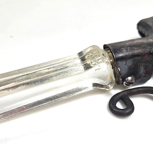 May include: A vintage bottle opener featuring a clear, rectangular acrylic handle attached to a dark, weathered metal head. The metal component includes a curved, hook-like lever mechanism secured with a small screw, showing signs of oxidation.