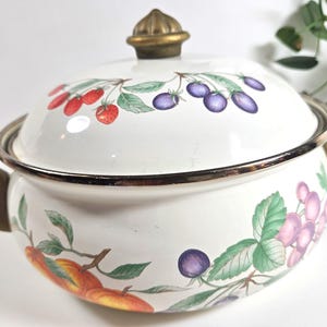 May include: A white enameled metal pot with a matching lid, featuring a colorful botanical print of strawberries, purple berries, and peaches with green leaves. The pot has two brass-toned metal side handles and a matching brass-toned lid knob.