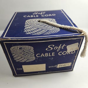 May include: A blue box with white lettering that reads "Soft Cable Cord" and "Color White". The box contains a spool of white cord.