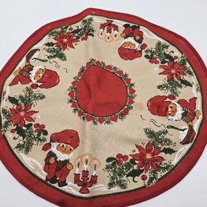 May include: A round Christmas tree skirt with a red border and a beige center. The skirt features illustrations of Santa Claus figures, poinsettias, and holly. The center has a solid red circle.
