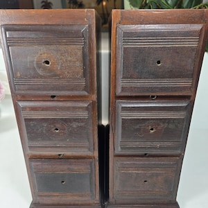 May include: Two tall, narrow wooden storage units, each featuring three stacked drawers with recessed rectangular panels and a single centered circular hole on each drawer front. The wood has a dark brown, weathered finish with visible grain.