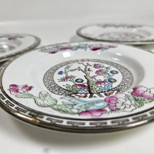 May include: Three white ceramic plates with a floral design. The plates feature a central motif of a tree and flowers in pink, blue, and green, surrounded by a decorative border. The rims have a gold and black geometric pattern.