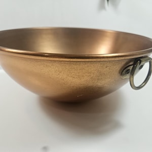 May include: A copper-colored bowl with a rounded shape and a slightly flared rim. The bowl has a handle on one side and a smooth, reflective surface. The bowl is likely used for mixing or serving food.