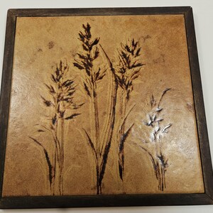 May include: Square wall art featuring a wheat field design. The artwork has a light brown background with dark brown wheat stalks. The artwork is framed in a dark brown wooden frame. The artwork is suitable for home decor.
